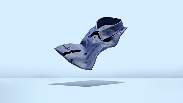 A chambray mens dress shirt floating weightlessly against a light blue background.
